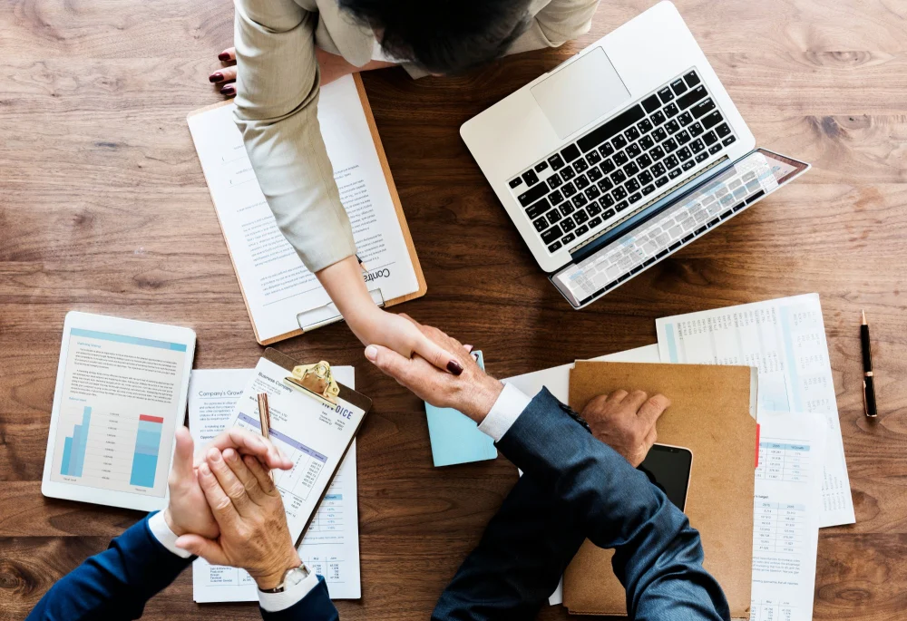Aperto de mão em negociação em inglês, com contrato, notebook e relatórios de negócios sobre a mesa